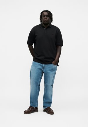 Mann mit dunkler Haut und Dreadlocks, trägt ein schwarzes Polohemd, hellblaue Jeans und braune Schuhe, steht mit den Händen in den Taschen vor weißem Hintergrund.