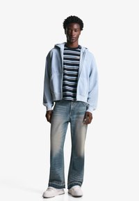 Helle blaue Zip-Hoodie mit Taschen, gestreiftes schwarz-weißes Shirt darunter, ausgebleichte blaue Jeans und weiße Sneakers.