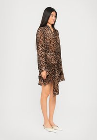 Donna con lunghi capelli neri che indossa un vestito leopardato a maniche lunghe e lunghezza al ginocchio, con delle décolleté bianche a punta e tacco basso, in piedi di lato su uno sfondo neutro.