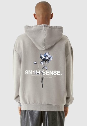 Person trägt grauen Kapuzenpullover mit blauem und weißem Blumenmotiv und dem Text "9N1M SENSE" auf dem Rücken, steht vor einem einfarbigen Hintergrund.