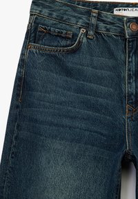 Jeans de mezclilla en azul oscuro con un acabado desgastado. Cuenta con cinco bolsillos, lazos para cinturón y costuras naranjas. Incluye un cierre de botón en la parte frontal.