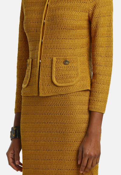 Nahaufnahme einer Person, die eine senfgelbe, strukturierte Strickjacke mit Fronttaschen und einen passenden Strickrock trägt.