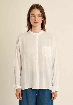 Jonge vrouw met lang bruin haar in een losse witte blouse met knopen en blauwe spijkerbroek, staand tegen een effen licht beige achtergrond.