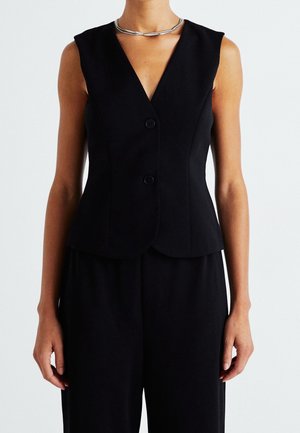 Kobieta w czarnej, bezrękawnikowej kamizelce na guziki z głębokim dekoltem w serek oraz luźnych czarnych spodniach, uzupełniona grubym srebrnym naszyjnikiem.