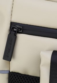 Krémová taška s texturovaným povrchem, ozdobená černým zipem a štítkem. Obsahuje síťovanou kapsu a šedý akcent.