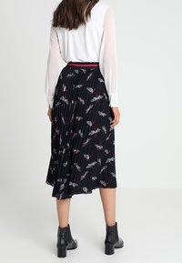 Gonna nera plissettata con stampa floreale, caratterizzata da accenti rossi e bianchi. Indossata con una blusa bianca e stivali neri alla caviglia, che mostrano il dettaglio della vita.