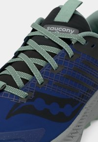 Suurendatud vaade sinisest Saucony tossust, millel on rohelised nöörid, must keel ja hallidega laiguline tald.