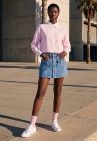 Jeune femme debout à l'extérieur, portant une chemise rose à manches longues, une jupe en jean à boutons sur le devant, des chaussettes roses et des baskets blanches, les mains dans les poches.
