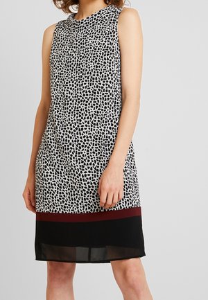 Ärmelloses knielanges Kleid mit unregelmäßigem schwarz-weißem Punktmuster, Burguindestreifen über einem einfarbigen schwarzen Saum, getragen von einer stehenden Person.