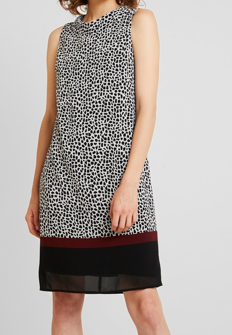 Robe sans manches jusqu'aux genoux avec un motif de points irréguliers noirs et blancs, une bande bordeaux au-dessus d'un ourlet noir uni, portée par une personne debout.