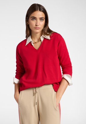 Donna con lunghi capelli castani indossa un maglione rosso a V sopra una camicia bianca con colletto e pantaloni beige con strisce rosse, mani nelle tasche.