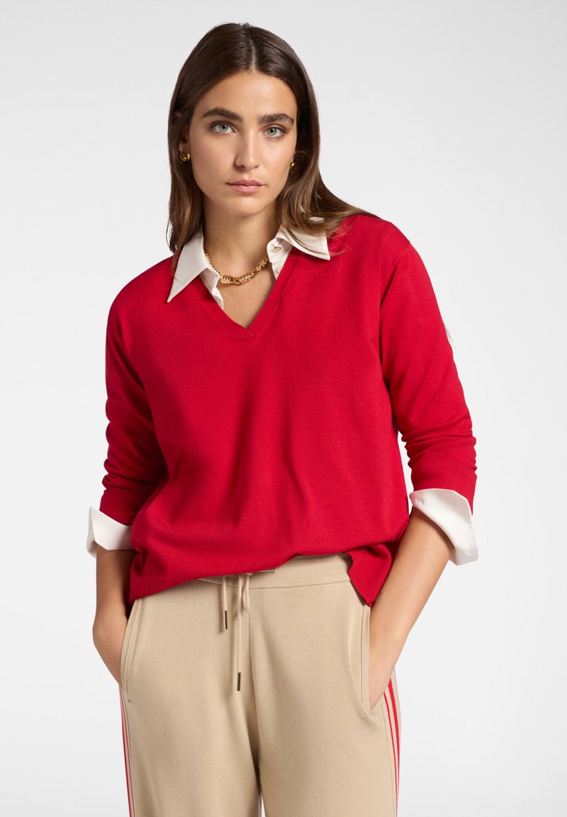 Nainen, jolla on pitkät ruskeat hiukset, käyttää punaista V-kaulaista neuletta valkoisen kauluspuseron päällä ja beigejä housuja, joissa on punaiset raidat, kädet taskuissa.