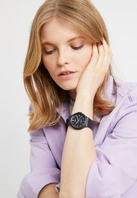Montre analogique noire avec un cadran rond et un design minimaliste. Bracelet en silicone et marquages clairs. Portée sur un poignet reposant contre le visage.