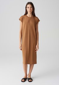 Braunes, kurzärmeliges Midikleid mit rundem Halsausschnitt und einer mittigen Naht. Glatte Textur, aus einem weichen Stoff hergestellt. Getragen mit schwarzen Slipper-Sandalen.