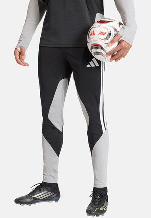 Persona con pantaloni sportivi neri e grigi e tacchetti che tiene una palla da calcio bianca e rossa nella mano destra.