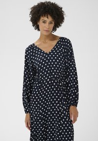 Robe marine à pois blancs, col en V, manches longues avec poignets froncés, et détail de ceinture nouée. Tissu lisse avec une coupe décontractée.