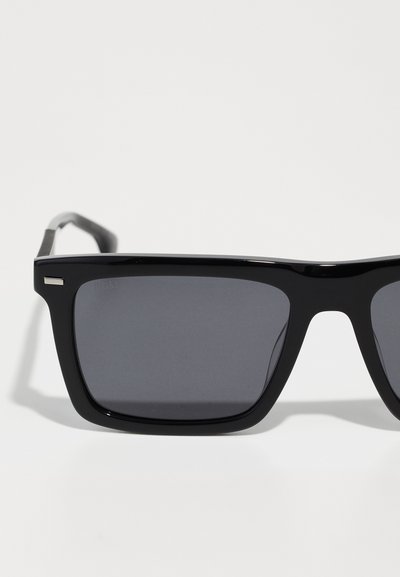 Lunettes noires avec une monture rectangulaire épaisse, des verres gris foncé et une finition brillante. Présente des accents métalliques fins sur les branches.