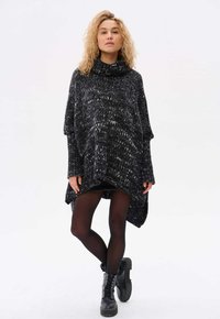 Czarny i szary dzianinowy poncho z wysokim kołnierzem, drapowanymi rękawami oraz luźnym krojem, w połączeniu z czarnymi rajstopami i butami wojskowymi.