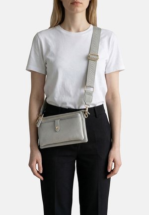 Donna che indossa una maglietta bianca e pantaloni neri con una borsa a tracolla grigio chiaro con trama, dotata di una tracolla larga regolabile e accessori dorati.