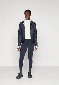 Lahka mornarsko modra vetrovka s sivimi poudarki in kapuco, nošena čez belo dolgoročno majico, kombinirana s prilegajočimi mornarsko moderimi pajkicami in supergami.