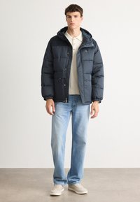 Νεαρός άντρας που στέκεται φορώντας ένα ναυτικό μπλε φουσκωτό μπουφάν με κουκούλα, ανοιχτόχρωμο μπλουζάκι πόλο, ανοιχτόχρωμα τζην και μπεζ αθλητικά παπούτσια, σε ουδέτερο φόντο.