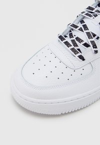 Λευκό δερμάτινο αθλητικό παπούτσι με διάτρητο μπροστινό μέρος, λαστιχένια εξωτερική σόλα και μαύρα κορδόνια με επαναλαμβαν όμενο μοτίβο κειμένου. Με υφή στο πάνω μέρος και ομαλές λεπτομέρειες.