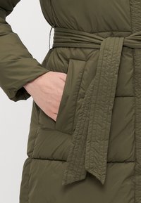 Olivengrøn pufferjakke med bælte i taljen, der har en sidelomme og en quiltet tekstur. Tøjet er glat og vandafvisende.