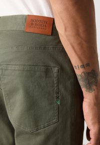 Pantalons vert olive avec un patch en cuir de marque, détail de poche arrière et broderie verte contrastante sur la couture de la poche.