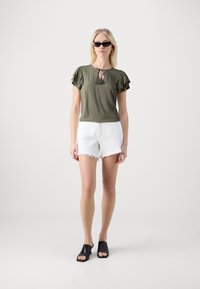 Oliven grønn topp med volangermed og nøkkelhullskrage, kombinert med hvite shorts med frynser og svarte åpen-tå mules. Glatte teksturer.
