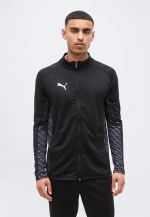 Vyras dėvintis juodą Puma striukę su užtrauktuku ir raštuotomis rankovėmis, stovintis atsisukęs į priekį priešais šviesią vientisą foną.