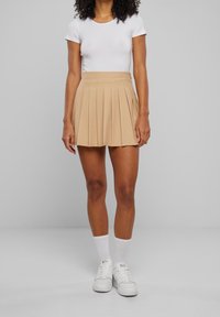 Mujer vistiendo una blusa blanca ajustada de manga corta, una falda corta plisada beige, calcetas blancas y zapatillas deportivas blancas, de pie contra un fondo gris liso.