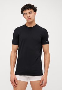 Camiseta negra de manga corta hecha de algodón, con un cuello redondo y un acento de logo en la manga izquierda. Textura suave, diseño ajustado.