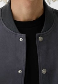 Gray suede bomber jacket with ribbed collar, snap buttons, and a smooth texture, layered over a black shirt.