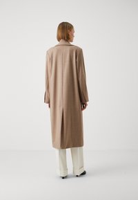 Cappotto lungo beige chiaro con colletto a punta, design su misura, aperture laterali e silhouette rilassata, abbinato a pantaloni crema e scarpe nere.