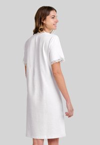Mujer de perfil vistiendo un vestido blanco hasta la rodilla con encaje en las mangas cortas y el escote, sonriendo frente a un fondo liso.