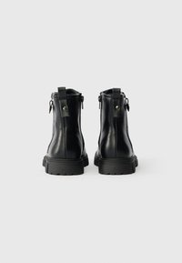 Par črne usnjene gležnjarje z debelimi gumijastimi podplati in zankami na hrbtni strani na osnovni beli ozadju.