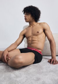 Shirtless man with curly hair seated on carpet, leaning on one arm, wearing black Tommy Hilfiger boxer briefs with red waistband.