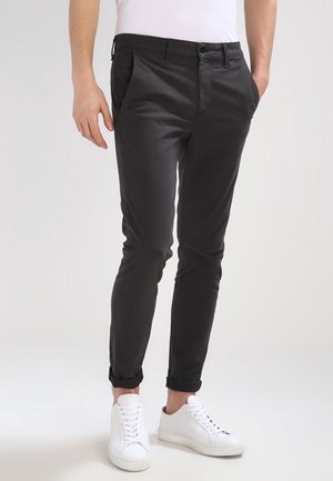 Mężczyzna w czarnych obcisłych spodniach z przednimi i tylnymi kieszeniami, w białych trampkach, stojący z rękami w kieszeniach.