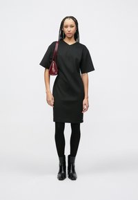 Μαύρο φόρεμα με κοντά μανίκια και V-λαιμόκοψη, από λείο ύφασμα, μήκους μέχρι το γόνατο, συνδυασμένο με μαύρες καλτσοδέτες και γυαλιστερά μαύρα μποτάκια μέχρι τον αστράγαλο· κρατάει μια μπορντό τσάντα.