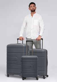 Uomo in camicia bianca che tiene le maniglie di tre valigie grigie a rotelle di diverse dimensioni su uno sfondo neutro.