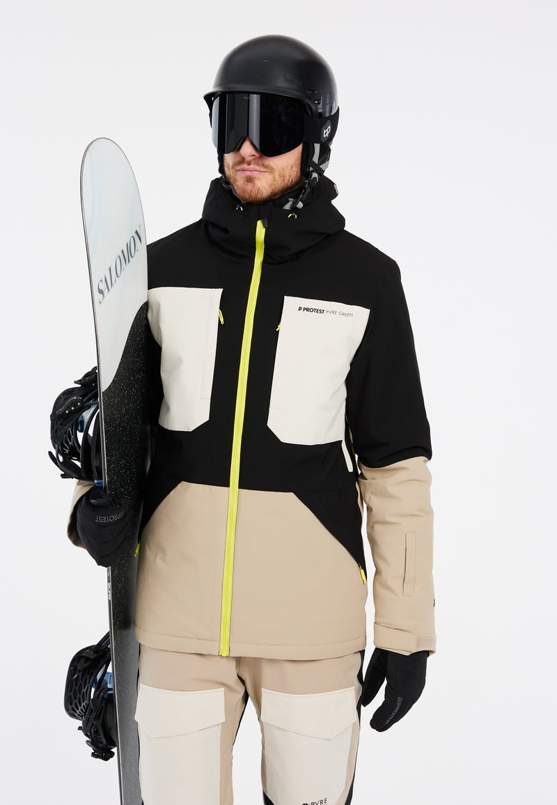 Snowboarder con giacca nera, beige e bianca con accenti gialli. Tiene una tavola da snowboard scura. I guanti sono neri e il casco è nero opaco.
