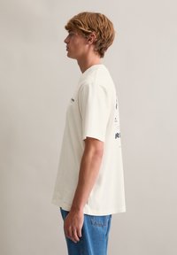 Korte mouwen, crèmekleurig katoenen T-shirt met zwarte bedrukte tekst op de achterkant en linkerborst, voorzien van een losse pasvorm en zijopeningen.