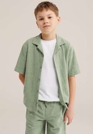 Rövid világosbarna hajú fiú texturált világoszöld gombos ingben és hozzá illő rövidnadrágban, egyszerű fehér póló felett, lazán áll.