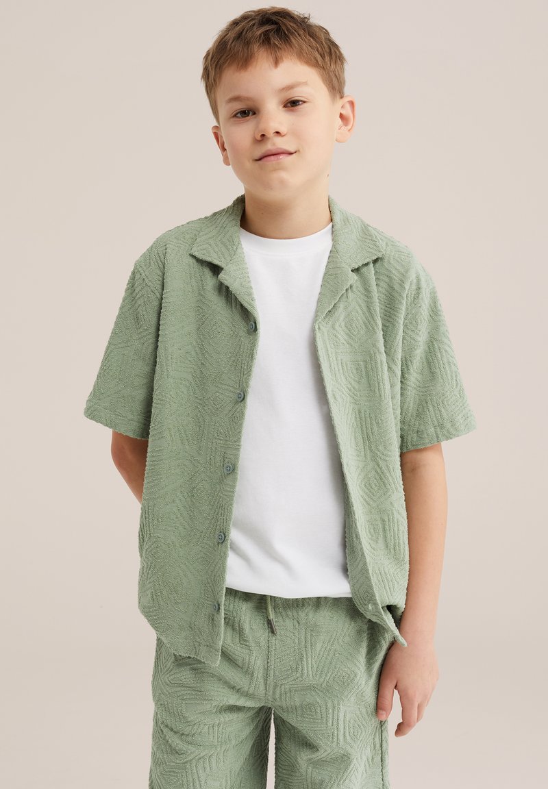 Boy with short light brown hair wears textured light green button-up shirt and matching shorts over a plain white T-shirt, standing casually.