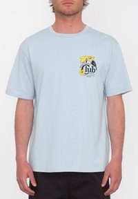 Camiseta de algodón de color azul claro con mangas cortas que presenta un estampado gráfico de flores amarillas y un pájaro, y el texto "Club" en negro.