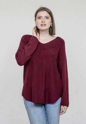 Jeune femme aux cheveux mi-longs portant un pull bordeaux et un jean bleu clair, touchant son cou, fond neutre.