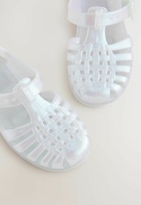Genomskinliga vita plastsandaler med ett vävt mönster, med stängd tå och justerbart ankelsnöre för en säker passform. Slät struktur.