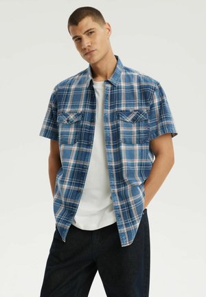 Mężczyzna o krótkich włosach, ubrany w niebieską koszulę w kratę z krótkim rękawem, na białej koszulce i ciemnych spodniach, stoi z rękami w kieszeniach.