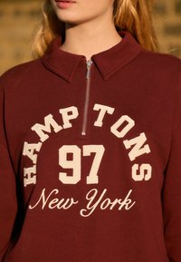 Tricou burgund cu guler înalt, fermoar argintiu și text brodat crem: "HAMPTONS 97 New York", având un material texturat.