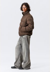 Brun pufferjakke med høy krage, med et quiltet design. Kombinert med løstsittende, lys grå denimjeans og svarte sko.